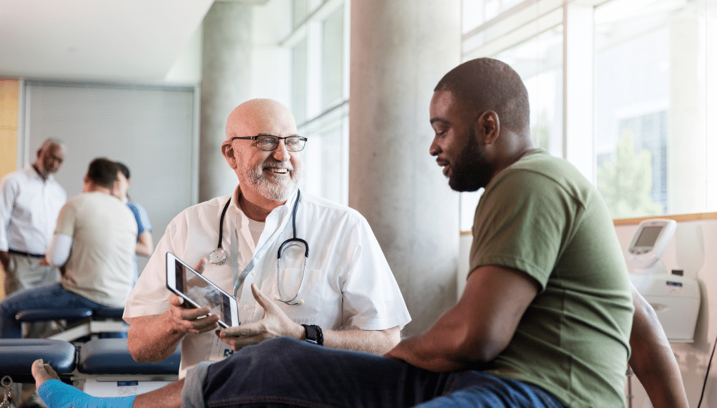 A doctor wearing a stethoscope shows a patient in a green shirt an image on his tablet.
