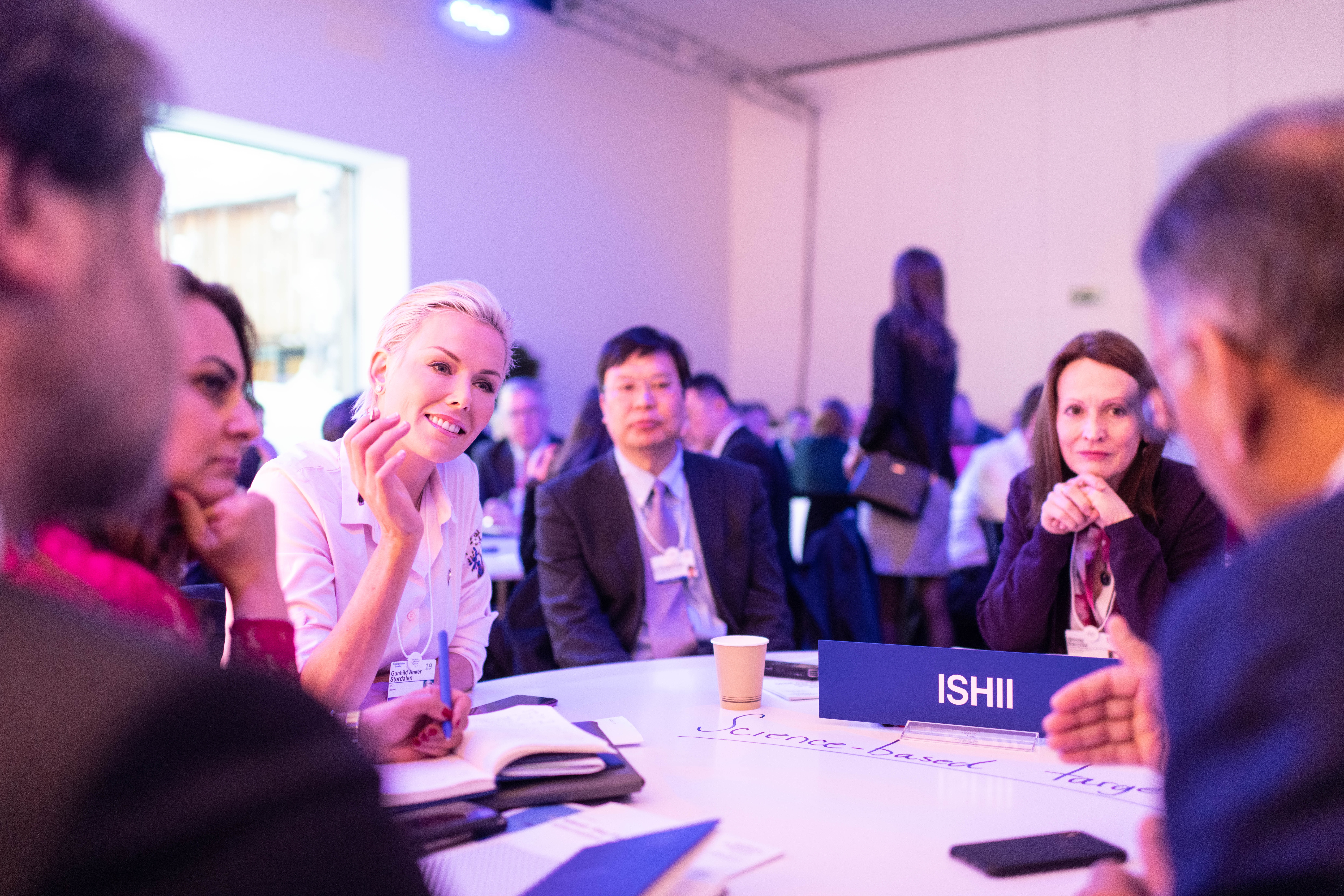 A group of people in professional dress clothes sit around a table at a conference.