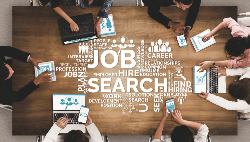 Seven people sit around a wooden table. The table has text describing job searches.