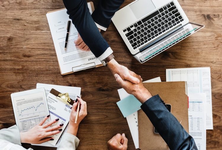 Two people shake hands over a wooden table with a laptop and papers on top of it.