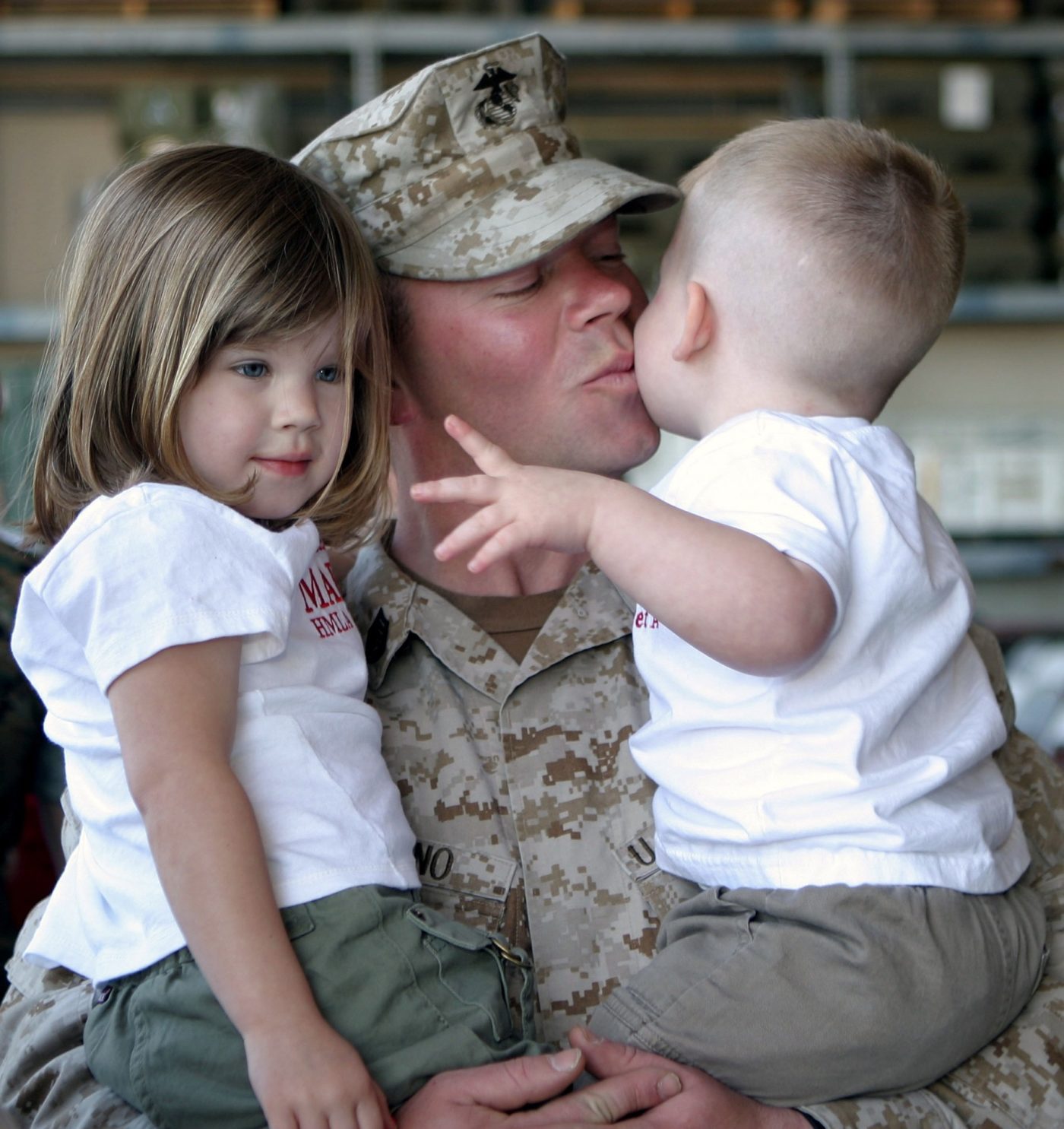 A man in a military uniform holds two children and kisses one of them on the cheek.