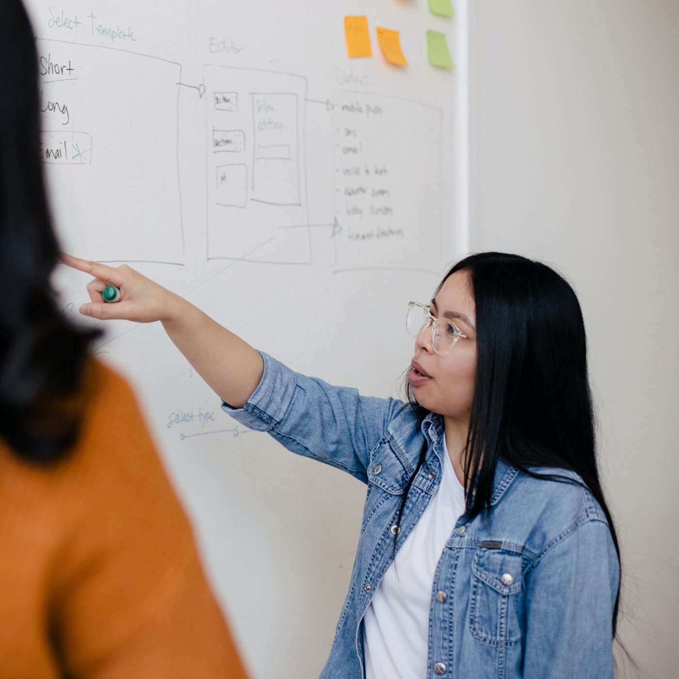 A woman in a jean jacket and glasses points to writing on a whiteboard.