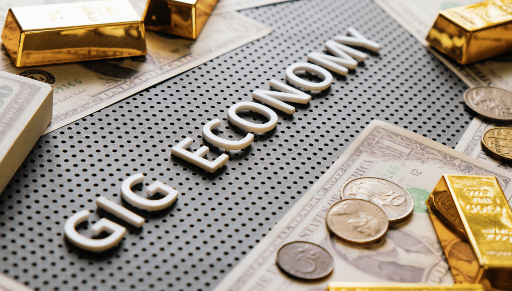 Some dollar bills, coins, and bars of gold lie on a table. In the middle of the table are the words "gig economy".