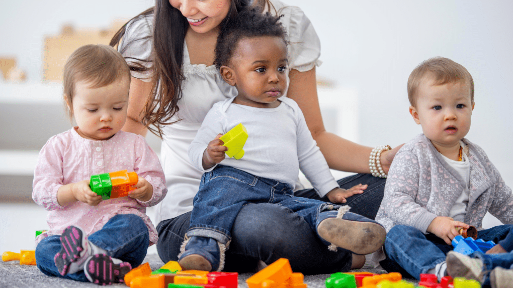 Three young children play with blocks. A woman sits behind them.