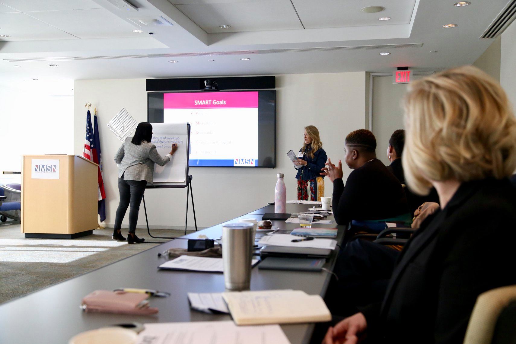 People sit at a long table covered in notebooks, facing a woman giving a presentation.