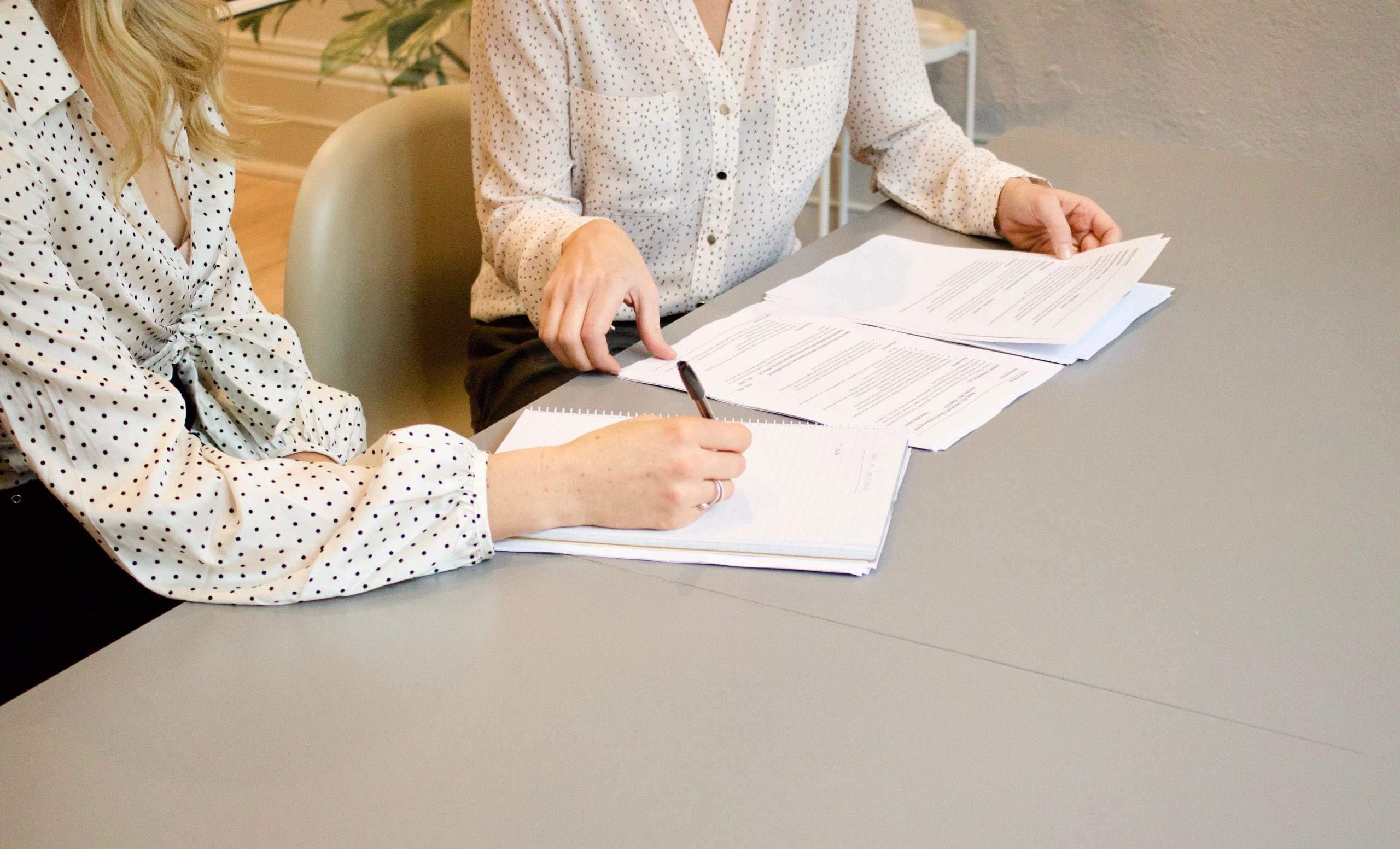 Two women sit at a gray table with papers on it.